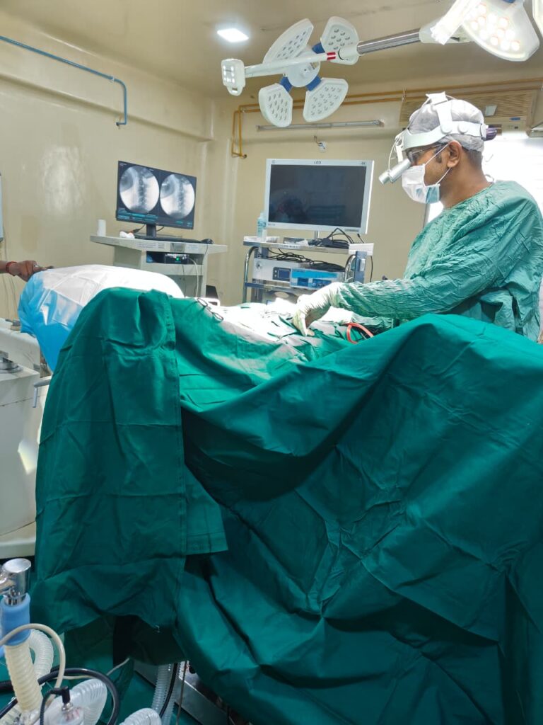 performs an operation on a patient A surgeon wearing green surgical scrubs, mask, performs an operation on a patient in a fully equipped operating room, with medical monitors and surgical lights visible overhead